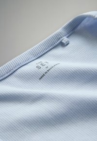 Hellblauer, gerippter Stoff mit strukturierter Oberfläche; in der Nähe des gerippten Halsausschnitts befindet sich ein Etikett mit der Aufschrift "THE SET M" und "MADE IN BANGLADESH".