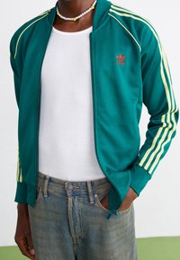 Grön sportjacka med gula stripes, dragkedja fram och röd logotyp. Bärs över en vit ribbad skjorta, tillsammans med blå denimjeans.