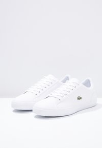 Paire de baskets blanches basses avec des lacets blancs et un logo crocodile vert sur le côté, posées sur une surface blanche.