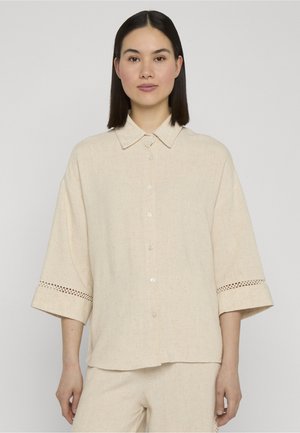 Frau mit glattem dunklem Haar, die ein lockeres beiges Hemd mit Knöpfen trägt, das weite Ärmel und gemusterte Aussparungen in der Nähe der Manschetten aufweist.