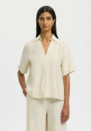 Femme aux longs cheveux noirs portant un haut crème à manches courtes avec col et un pantalon large assorti, sur un fond clair uni.