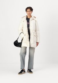 Jachetă puffer albă matlasată, cu glugă, închidere cu fermoar și capse, buzunare frontale; asociată cu un tricou cu grafică, blugi deschiși și adidași negri.