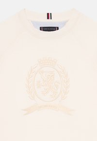 Krämfärgad sweatshirt med ett broderat emblem med ett lejon, omgivet av lagerblad och en banner, med "MCMLXXXV" inskrivet.