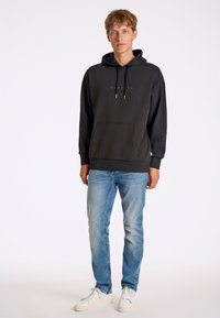 Sudadera negra con bolsillo tipo canguro, capucha con cordón y detalle de logo. Combinada con jeans de mezclilla clara y zapatillas blancas. Diseño simple y casual.