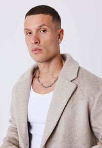 Cappotto grigio chiaro in misto lana con colletto a bavero, abbinato a un top bianco aderente. Caratterizzato da una collana con catena d'argento e orecchini a perno con diamanti.