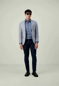 Man wearing sunglasses, light gray blazer, blue button-up shirt, dark blue pants, and black shoes standing against plain background.