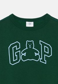 Dunkelgrünes Baumwoll-T-Shirt mit einem weißen Umriss eines Bären und den Buchstaben "G" und "P". Ribbeliger Rundhalsausschnitt mit weicher Textur.