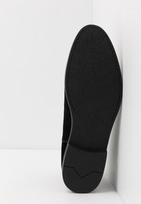 Zapato de ante negro con una suela de goma negra texturizada, punta ligeramente puntiaguda y un diseño angular distintivo en el talón.