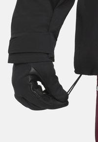 Schwarze Handschuhe aus weichem Stoff und Leder, mit einer strukturierten Grifffläche und einer Zugverstellung für einen sicheren Sitz am Handgelenk.