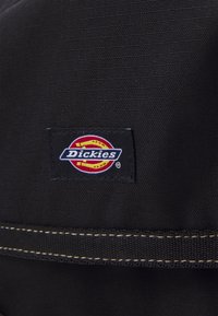 Μαύρο ύφασμα με υφή διαθέτει ένα ορθογώνιο λογότυπο Dickies σε κόκκινο, κίτρινο και μπλε χρώμα, με λευκές ραφές.