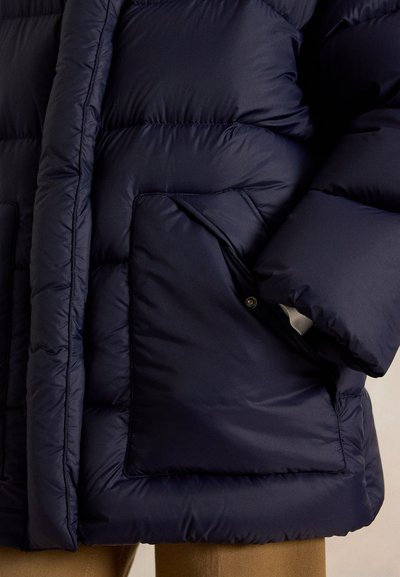 Doudoune matelassée bleu marine foncé avec grande poche à pression portée sur un pantalon marron clair.
