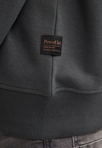 Sudadera gris oscuro con un dobladillo acanalado y una etiqueta cosida con "Petrol Ind" en naranja. Tejido suave con una textura lisa.