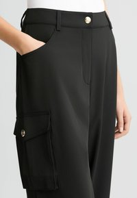 Zwarte broek van gladde stof, voorzien van zijzakken en een cargozak met een gouden knoop detail, nauwsluitende pasvorm met rechte pijpen.