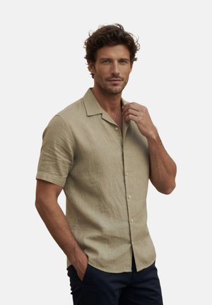 Hombre vistiendo una camisa beige de manga corta con cuello y botones, una mano ajusta el cuello, la otra mano en el bolsillo, sobre un fondo liso.