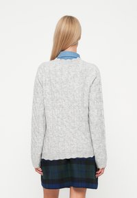 Grå kabelstrik sweater med lange ærmer, der har en bølget halsudskæring. Båret over en blå ternet nederdel. Blød tekstur.