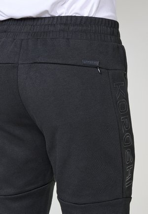 Tuva melnā sporta bikses ar elastīgu jostu un "KOROSHI" logo, kā arī ar kabatām ar rāvējslēdzējiem sānos un aizmugurē.