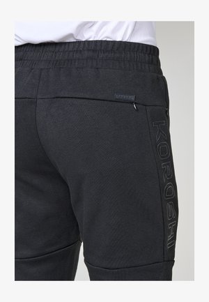 Nahaufnahme von schwarzen Jogginghosen mit elastischem Bund, die das "KOROSHI"-Logo und Reißverschlusstaschen an den Seiten und hinten zeigen.