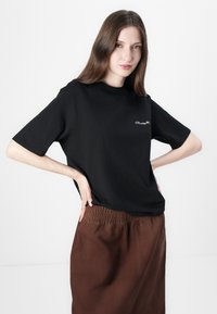 T-shirt nera a maniche corte in cotone con scollatura rotonda e piccolo testo ricamato bianco sul petto, abbinata a una gonna marrone con vita elasticizzata.