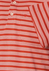 Czerwono-różowa polo w paski z kołnierzykiem na dwa guziki, teksturowana tkanina i mała kieszeń na piersi. Akcentowana kontrastującymi guzikami.