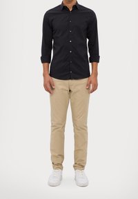 Tmavá košile na knoflíky s dlouhými rukávy, kombinovaná s béžovými chinos kalhotami a bílými teniskami. Má slim fit střih a hladkou texturu látky.