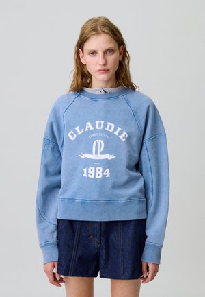 Sudadera de algodón azul claro con mangas raglán, que presenta un logo blanco y el año "1984". Combinada con pantalones cortos de mezclilla oscura, diseño simple.