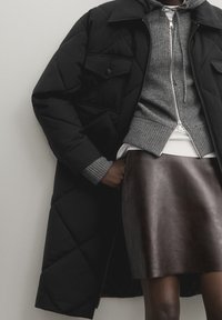 Persoon in een zwarte gewatteerde jas over een grijze rits-hoodie en een donkerbruine leren rok, staande tegen een lichte muur.