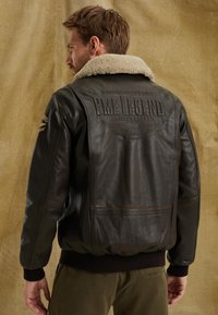 Brun läderjacka med shearling-krage, präglad logotyp på baksidan, ribbade ärmslut och nederkant, med sidofickor och kontrasterande sömmar.