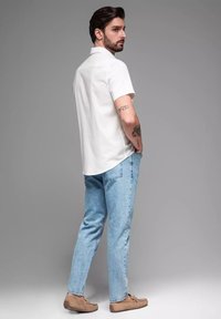 Witte kortemouwen shirt, lichtblauwe spijkerbroek en bruine instapschoenen. Man staat met handen in de zakken, zichtbare tattoos op zijn arm.