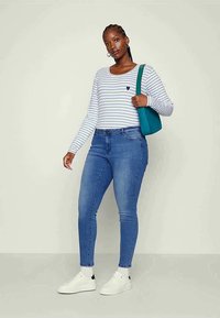 Long-sleeve, white and blue striped top with small heart, paired with blue skinny jeans. Model wears white sneakers and holds a teal bag.