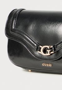 Black faux leather handbag with a curved flap, gold-tone hardware, embossed logo, and subtle stitching details. Compact design.