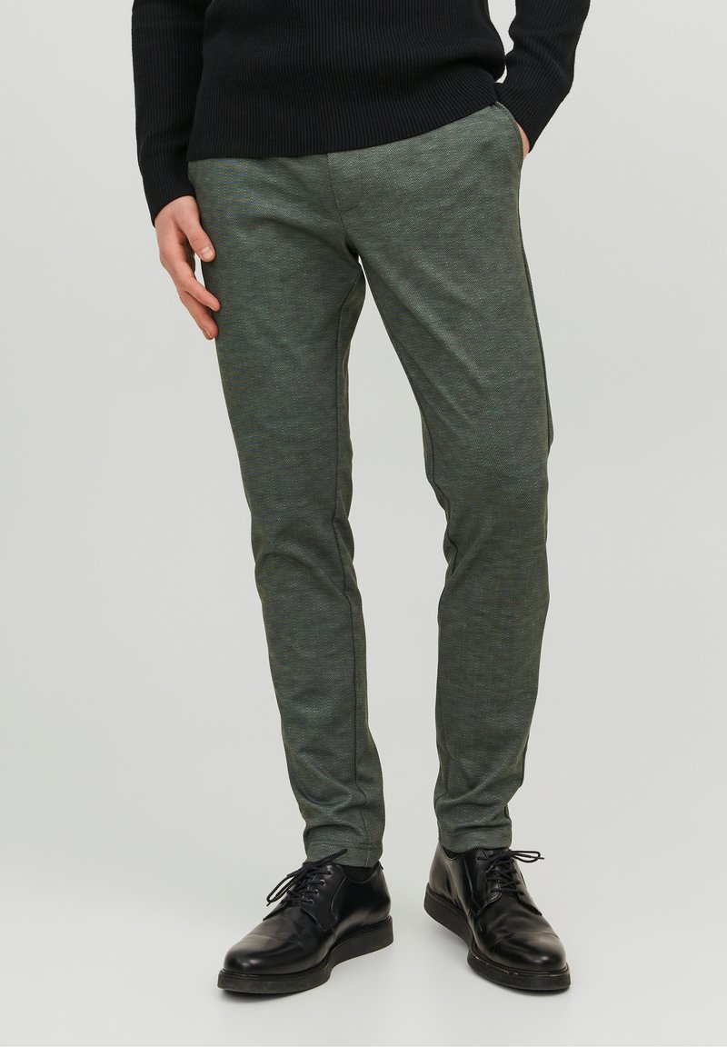 Man in een slim-fit grijze broek met zwarte zijstreep en zwarte schoenen, tegen een effen lichte achtergrond.