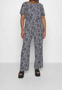 Marinblå jumpsuit med vit virvelmönster; korta ärmar, lös passform, vida ben; bärs med svarta plattformssandaler. Texturerad tyg, avslappnad design.