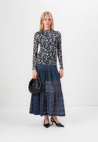 Long-sleeve top in dark floral mesh with high neckline, paired with a tiered navy lace skirt and black handbag; pointed-toe heels.