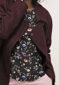 Cappotto in lana bordeaux con chiusura a bottoni sopra un top a fantasia floreale nera con fiori rossi, blu e crema su uno sfondo scuro.