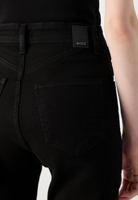 Jeans en denim noir avec un design standard à cinq poches, incluant une poche arrière carrée et une étiquette de marque en cuir noir sur la ceinture.