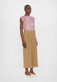 Sleeveless top in light pink with floral patterns, paired with beige wide-leg trousers. Model wears white heeled mules, showcasing a clean silhouette.
