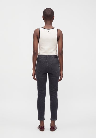 Personne debout de dos portant un haut blanc sans manches avec bordure noire, un jean slim gris foncé et des chaussures rouge foncé sur fond uni.