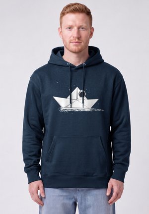 Mann mit roten Haaren trägt einen dunkelblauen Hoodie mit einem weißen Papierboot, auf dem ein Astronaut auf dem Wasser treibt.