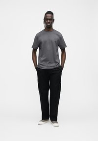 Ciemnoszary t-shirt z krótkim rękawem i subtelnym logo, zestawiony z czarnymi spodniami cargo z bocznymi kieszeniami oraz jasnoszarymi sneakersami.