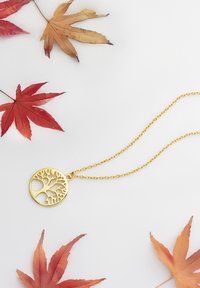 Goldener Lebensbaum-Anhänger an einer zarten Kette, mit ausgeschnittenen Zweigen und Blättern, vor herbstlich gefärbten Ahornblättern.