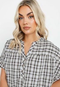 Blusa a maniche corte con bottoni, in pattern a quadri grigi e bianchi, caratterizzata da un colletto arrotondato e dettagli arricciati alle spalle.