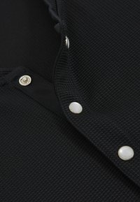 Detail blízkého pohledu na černou texturovanou látku se stříbrnými kovovými knoflíky a detaily s cvočkovým zapínáním podél výstřihu.