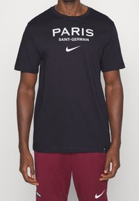 Czarna bawełniana koszulka z nadrukiem „PARIS SAINT-GERMAIN” w białym kolorze, ozdobiona logo Nike na przodzie. Okrągły dekolt, krótkie rękawy.
