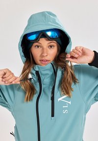 Light blue ski jacket with a high collar, black accents, two zippered pockets, adjustable hood, and a white logo on the front.