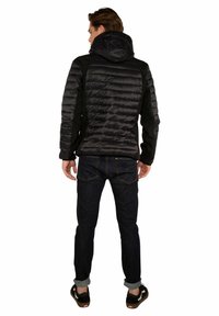Giacca nera imbottita con cappuccio, caratterizzata da una texture trapuntata sulla schiena, abbinata a jeans scuri e calzature casual.