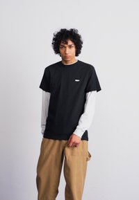 Μαύρο κοντομάνικο μπλουζάκι πάνω από ένα λευκό μακρυμάνικο πουκάμισο, συνδυασμένο με μπεζ cargo παντελόνι. Απλός σχεδιασμός, χωρίς ορατά μοτίβα ή διακοσμήσεις.