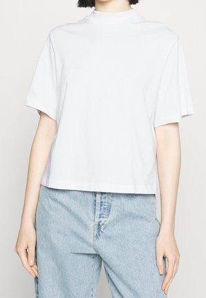 Person trägt ein schlichtes, weißes Kurzarmshirt mit Stehkragen und hellblaue, hoch taillierte Jeans vor neutralem Hintergrund.