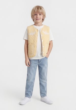 Blond chlapec v bielom tričku, žltej prešívanej viestke s vtáčími motívmi, svetlomodrých džínsoch a bielych teniskách, stojaci s jednou rukou vrecku.