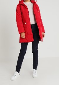Veste matelassée rouge avec capuche, fermeture éclair à l'avant et poches latérales. Portée sur un pull léger et associée à un pantalon bleu marine et des baskets blanches.