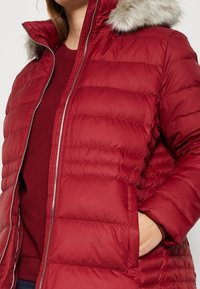 Vrouw in een rode gewatteerde jas met een capuchon afgewerkt met grijs bont, hand in zijzak, over een rode trui.